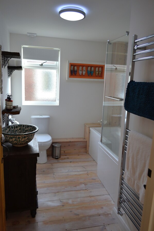 Une salle de bain avec un plancher en bois, des toilettes blanches, un lavabo blanc agrémenté d’un récipient décoratif, une cabine de douche en verre et un porte-serviettes.
