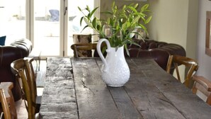 Een rustieke eettafel met een witte kan groene planten, omringd door houten stoelen.