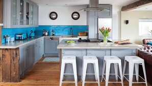 Een moderne keuken met blauwe kastjes, een centraal eiland met krukken en een houten plafond, met uitzicht op de woonkamer.