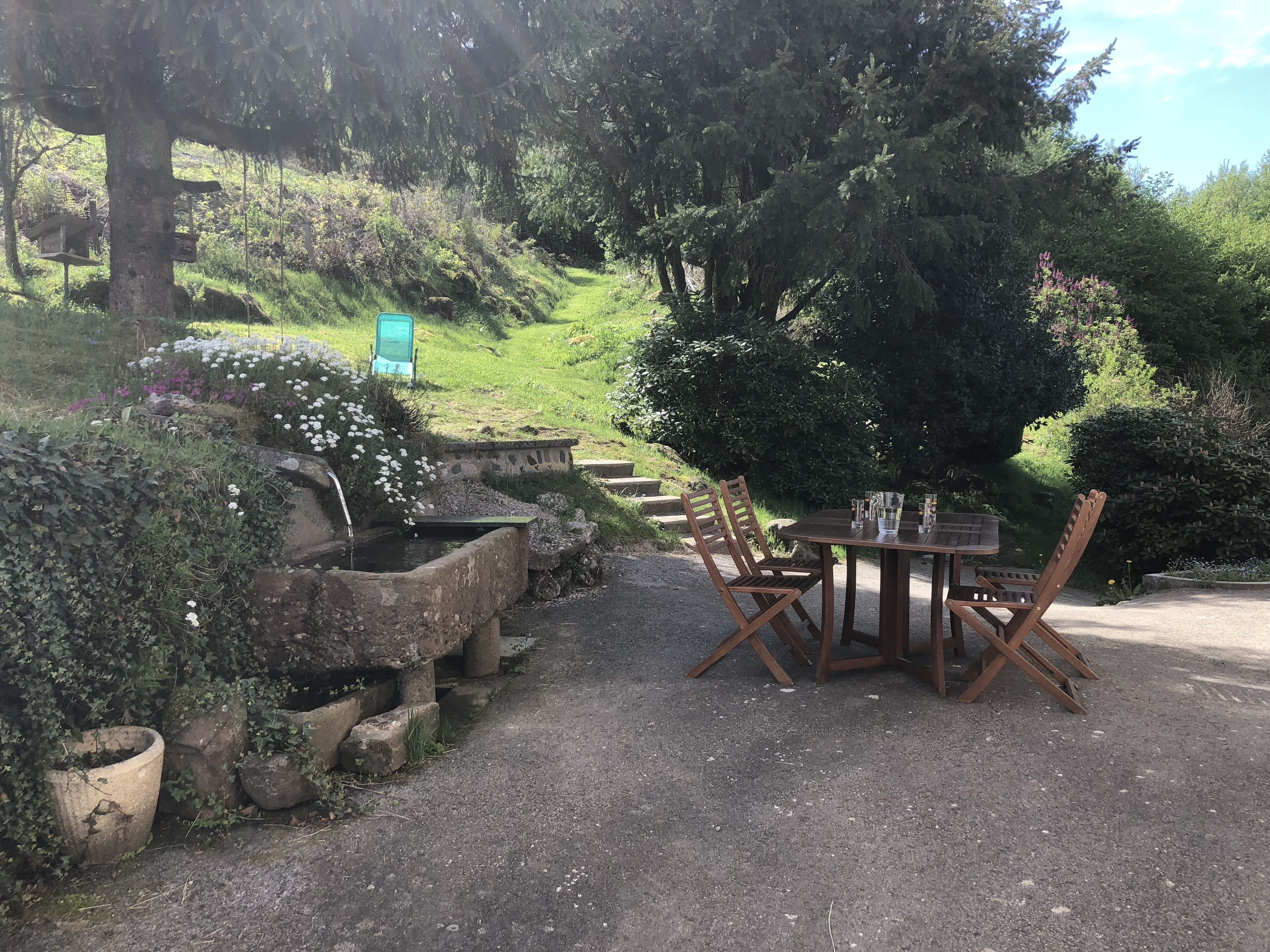A rustic outdoor seating area with a stone table and wooden chairs, surrounded by lush greenery and a small pond.