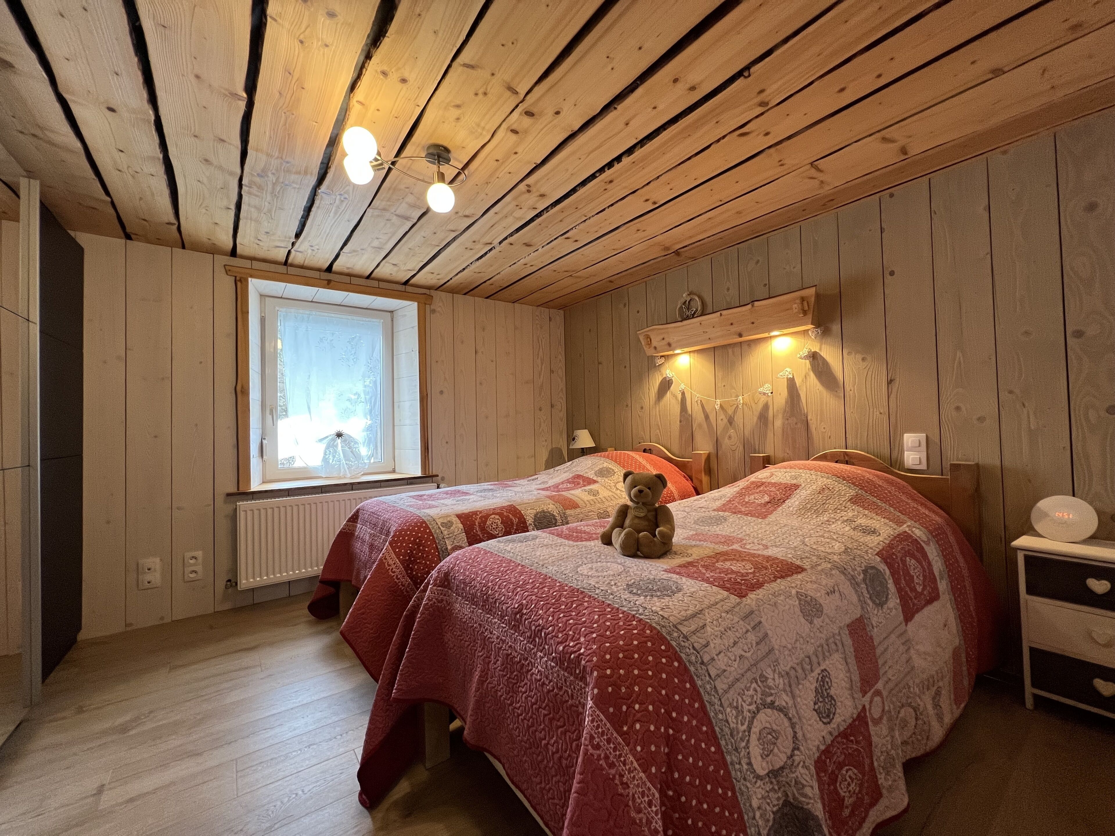 A cozy bedroom with wooden walls and ceiling, two beds with red and white bedding, a window with curtains, and a nightstand with a clock.