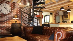 A cozy room with a brown leather sofa, a wooden table, a brick wall, a spiral staircase, and a kitchen in the background.