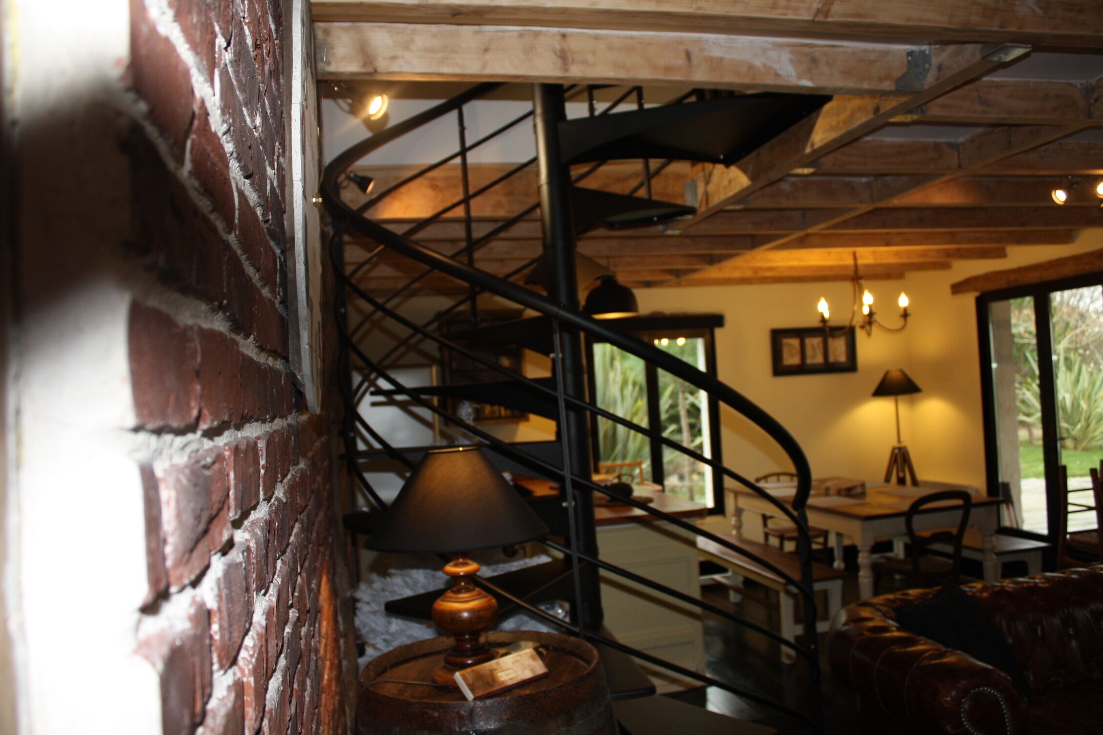 A rustic interior with exposed brick walls, a spiral staircase, a leather sofa, and a table with a lamp.