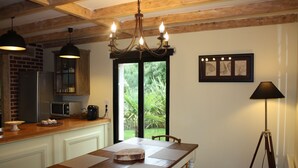 A dining area with a wooden table, placemats, and a chandelier. There is a kitchen area with a refrigerator, microwave, and cabinets. A window provides a view of greenery outside.