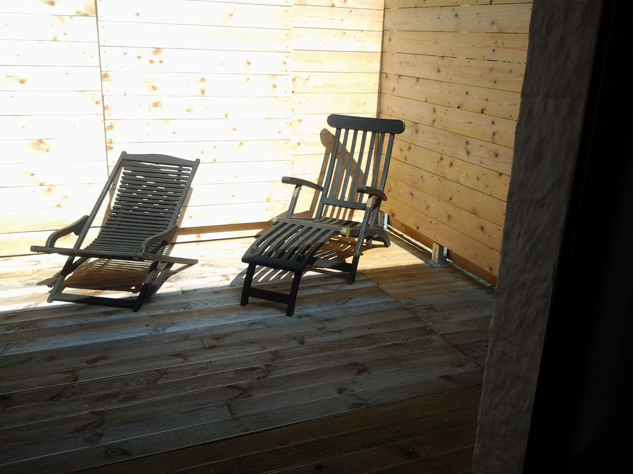 Two wooden deck chairs with metal frames, positioned on a wooden deck with a wooden wall in the background.