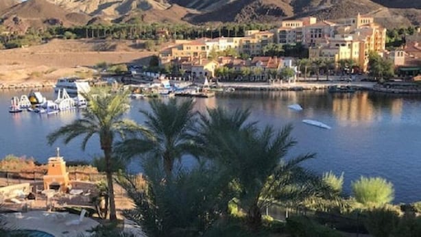 Un pueblo costero con un puerto deportivo, palmeras y montañas al fondo.