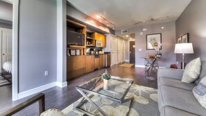 Una sala de estar moderna con una mesa de centro de vidrio, un sofá gris y una zona de cocina con armarios de madera y un microondas.