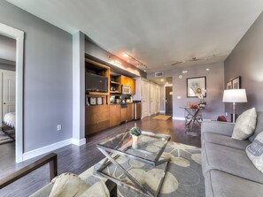 Un salon moderne avec une table basse en verre, un canapé gris et un coin cuisine doté de meubles en bois et d’un four à micro-ondes.