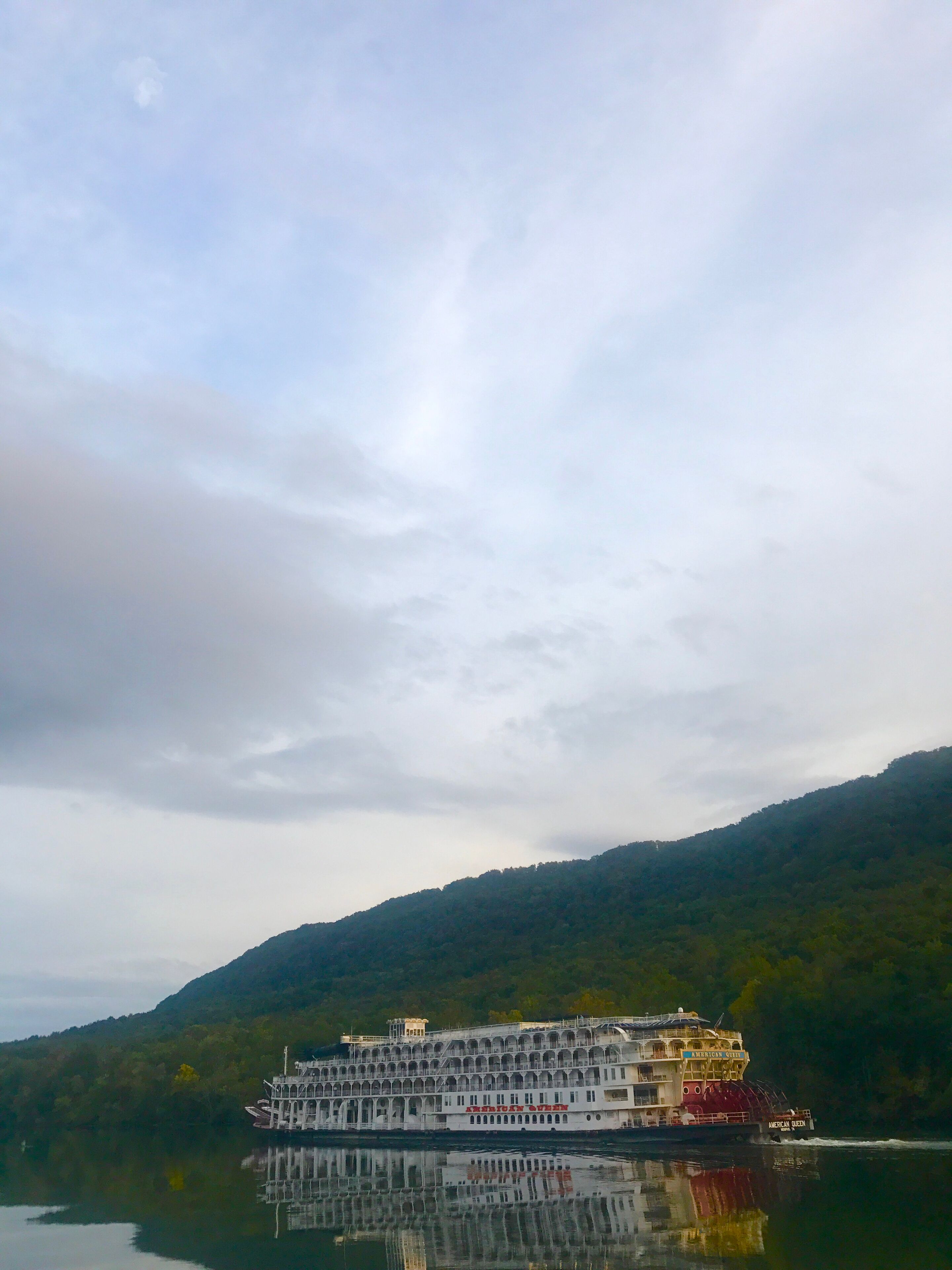 A large riverboat with multiple decks and a covered upper deck, docked at a calm river with a forested hillside in the background.