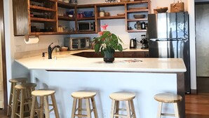 A modern kitchen with a central island, wooden stools, built-in shelves, and a stainless steel refrigerator. - Private beach front property, the perfect getaway! Enjoy our slice of paradise. (Waimea)