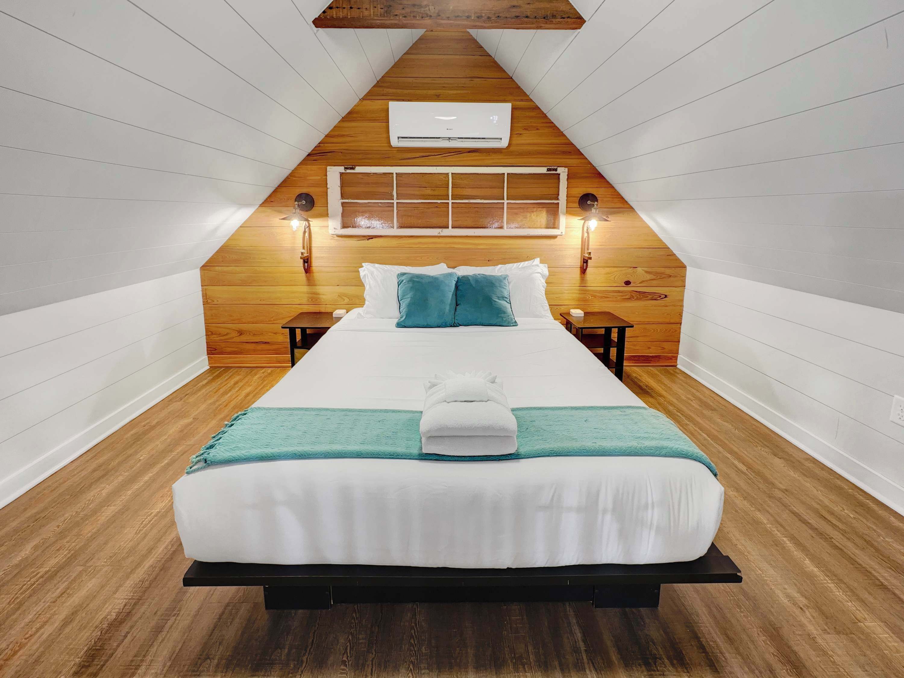 Ein Schlafzimmer mit schrägen Wänden, einem Bett mit weißer Bettwäsche, einer türkisfarbenen Tagesdecke und zwei Nachttischlampen.