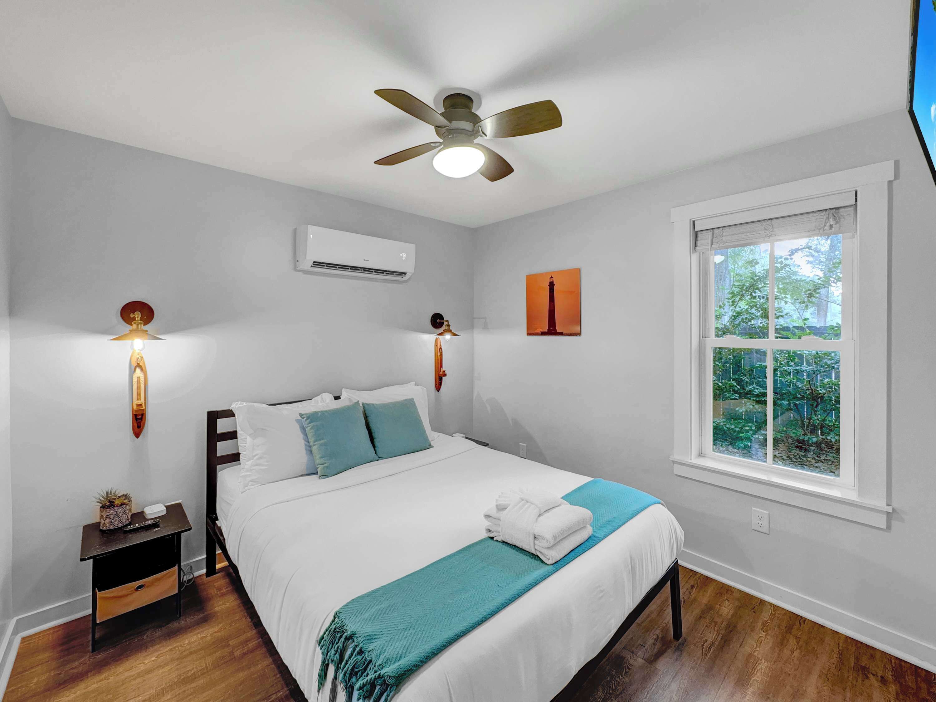 Une chambre à coucher comprenant un lit, un ventilateur de plafond, un appareil de climatisation, une table de chevet, une lampe fixée au mur et une fenêtre donnant sur un paysage verdoyant.