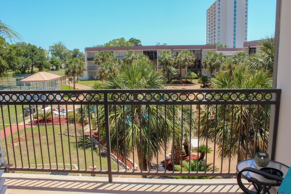 A balcony with a metal railing, overlooking a courtyard with palm trees and a building in the background.