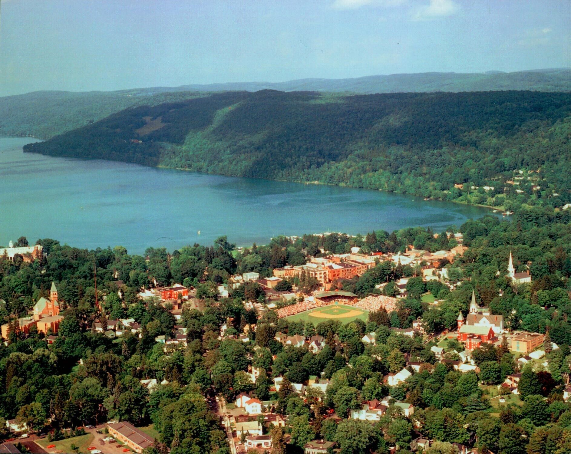 Vista aérea de un pueblo con un lago, rodeado de bosques densos y colinas.