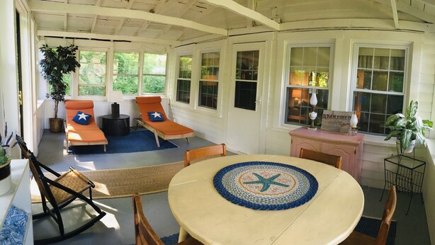 A cozy, sunlit room with a round table, two orange chairs, a rocking chair, and a small desk with a sign. - StarField Cottage...The Perfect Cooperstown Dreams Park Getaway! (Cooperstown)