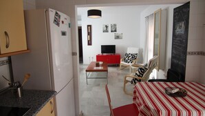 Ein kompakter Wohnraum mit Küchenzeile, einem Esstisch mit rot-weiß gestreifter Tischdecke, einer Couch mit gemusterten Kissen, einem Fernseher und einer Tafel mit Schriftzügen an der Wand.