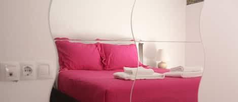 Reflejo de una cama con ropa de cama rosa, una lámpara de noche y toallas blancas dobladas con cuidado.