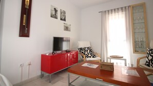 Ein Hotelzimmer mit einem roten Schrank, einem Fernseher, einem Sessel mit gemustertem Kissen, einem kleinen Tisch und einem Fenster mit Vorhängen.