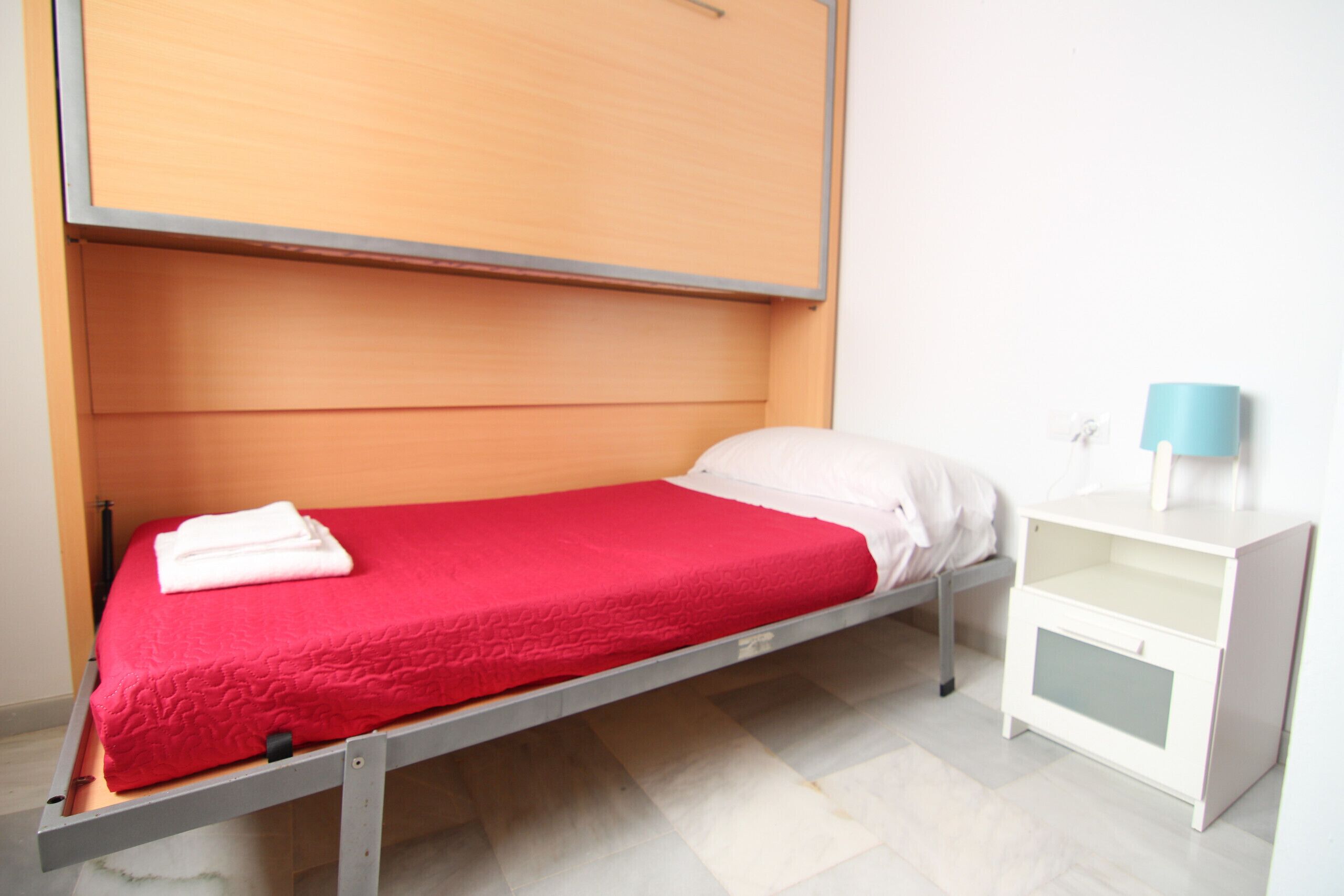 Ein Einzelbett mit roter Matratze und weißen Kissen, ein Nachttisch in Weiß mit Lampe sowie ein Holzschrank über dem Bett.