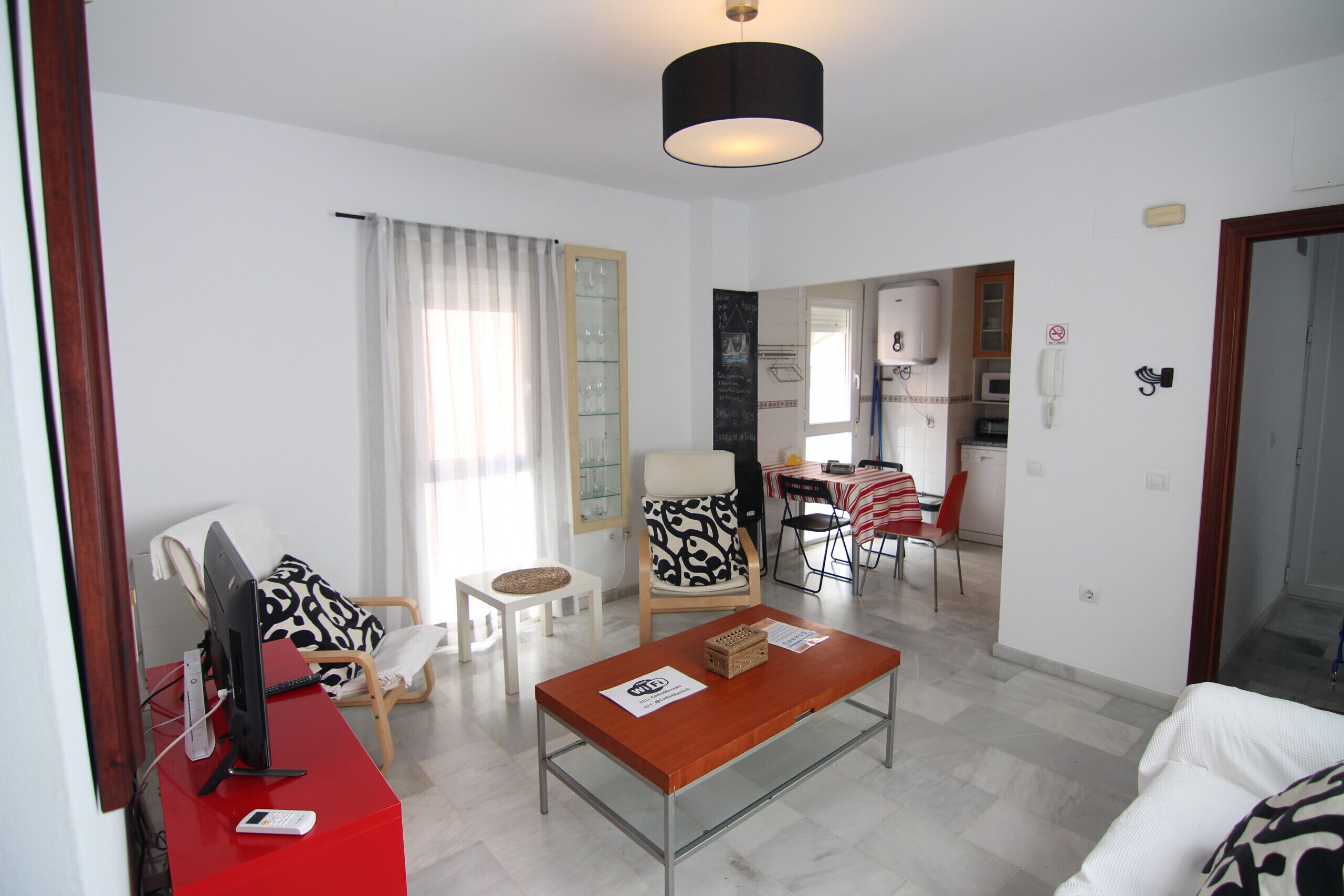 Ein modernes Wohnzimmer mit einem roten Schreibtisch, einem weißen Sofa und einem gemusterten Sessel. Es gibt einen Essbereich mit Tisch und Stühlen, und im Hintergrund eine Küche.