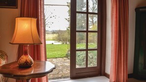 Ein Zimmer mit einer Tischlampe, einem Fenster mit Vorhängen und Blick auf einen Garten.