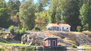 En stuga med röd fasad och vita detaljer, belägen på en klippig strand vid en sjö.