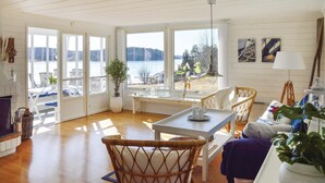 Ett rymligt rum med ett stort fönster med utsikt över en sjö, en matplats med flätade stolar och en mysig sittgrupp med en soffa och en lampa.