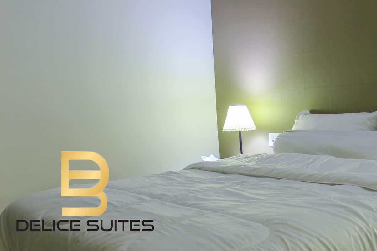 A neatly made bed with white bedding, two bedside lamps, and a logo for Delice Suites in the bottom left corner.