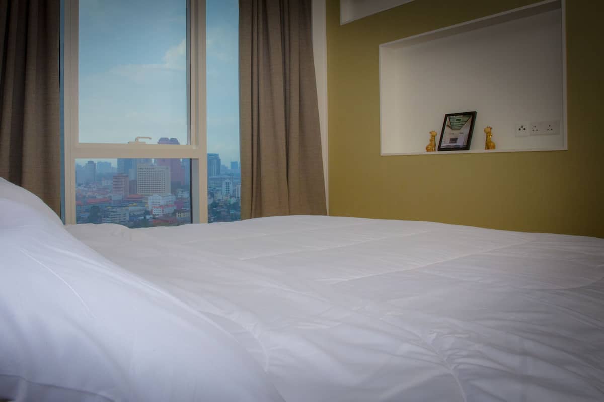 A hotel room with a large bed, a city view through a window, and a shelf with a picture and two small figurines.