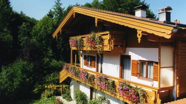 A traditional chalet with a wooden balcony adorned with flowers, set against a backdrop of dense forest. - Ferienwohnung, 55 qm, 2 Schlafzimmer, Balkon und Terrasse (Siegsdorf)