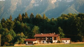 A two-story house with a red roof, surrounded by a forest and mountains in the background. - Ferienwohnung, 55 qm, 2 Schlafzimmer, Balkon und Terrasse (Siegsdorf)