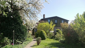 En sti gennem en have fører til et hus med skorsten, omgivet af frodig vegetation og blomstrende planter.