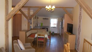 En hyggelig stue med sofa og spisebord, træbjælker, en lysekrone og et køkken i baggrunden.