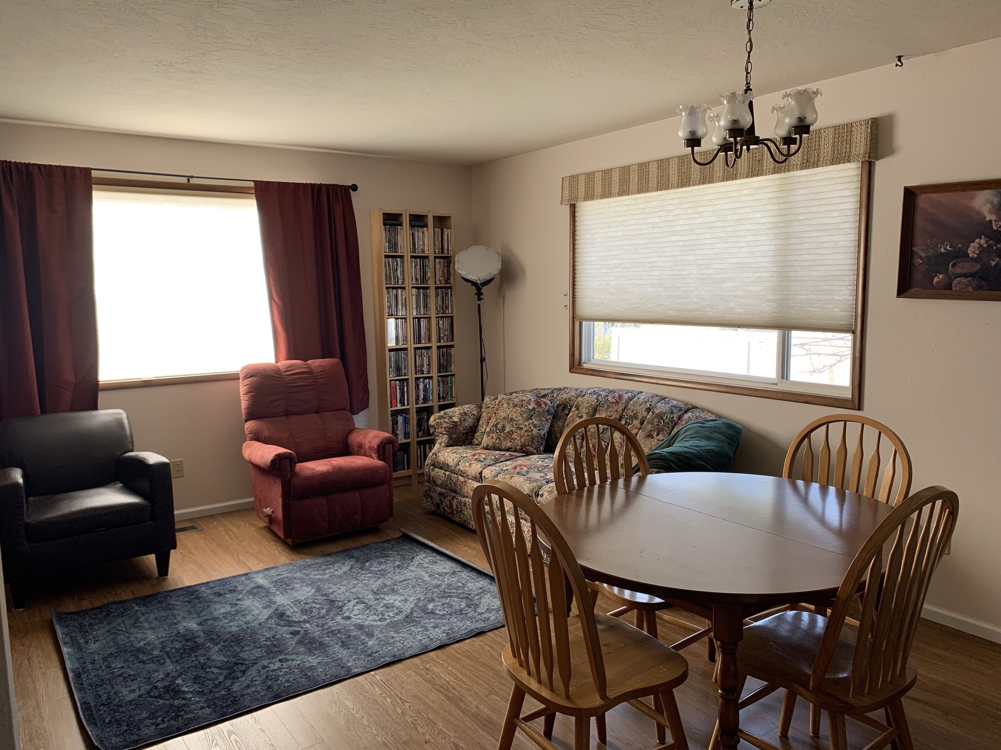 A living room with a sofa, a red armchair, a dining table with chairs, a bookshelf, and a window with blinds.