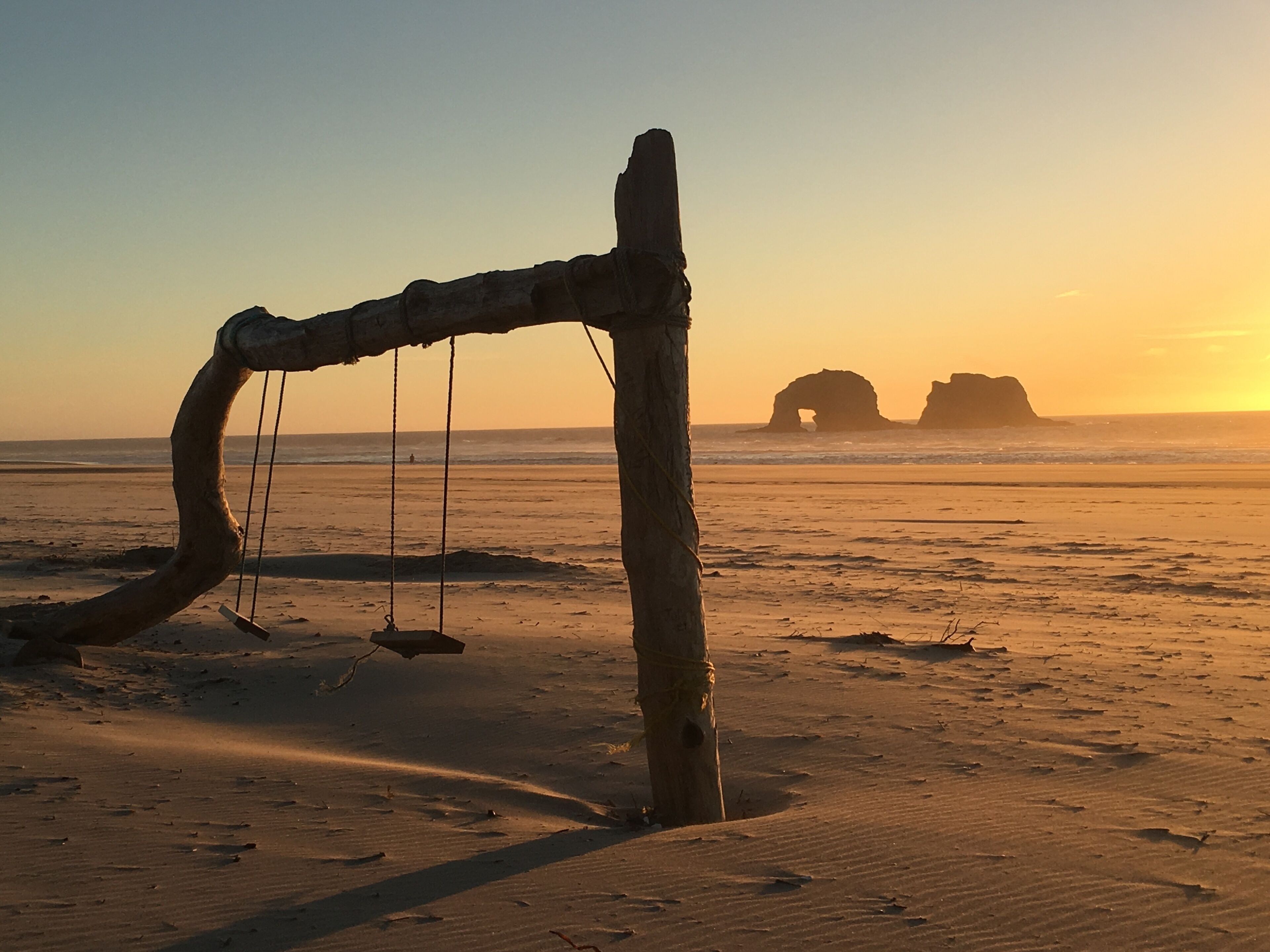 A wooden swing set on a sandy beach with a sunset in the background.