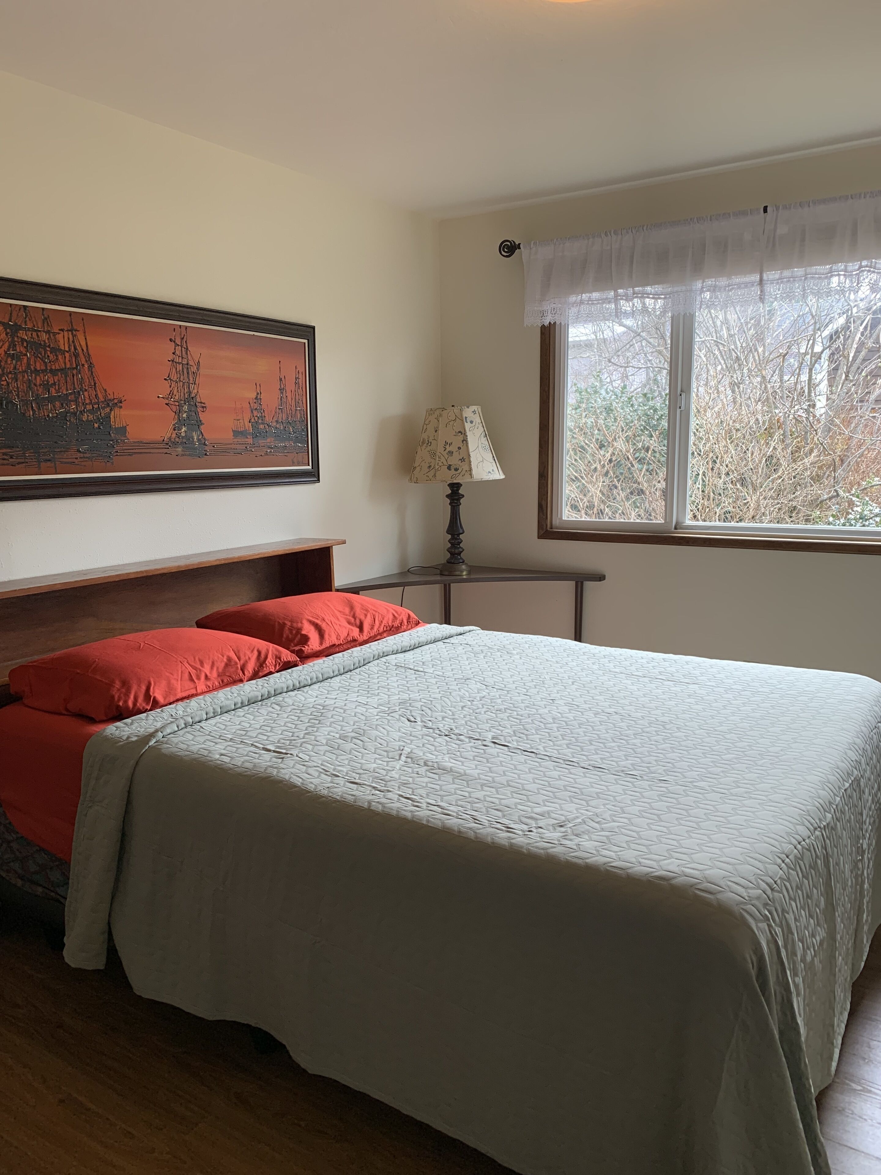 Un dormitorio con una cama grande, una mesita de noche, una lámpara, una ventana con vistas a árboles y un cuadro en la pared.