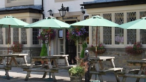Outdoor seating with green umbrellas and stone tables in front of a building with large windows and hanging flower baskets. - The Miners Arms (Hope Valley)