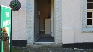 A building entrance with a door, a hanging flower basket, and a green sign with white text. - The Miners Arms (Hope Valley)