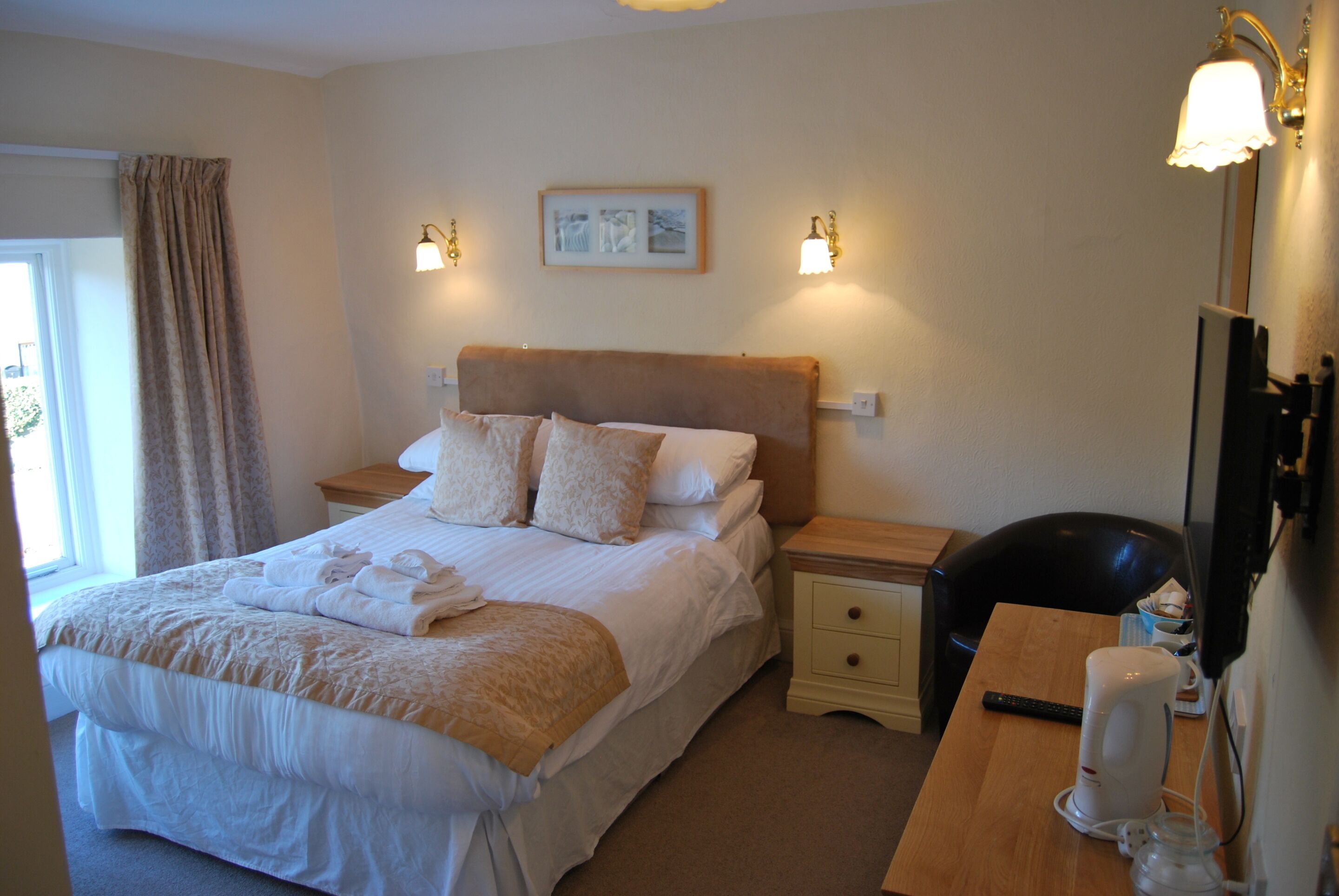 A neatly made bed with pillows, a bedside table with a lamp, a chair, and a desk with a cup and remote control.