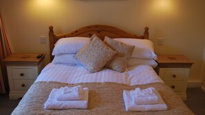 A neatly made bed with pillows, a blanket, and folded towels. There are bedside tables with a remote control and a book on one of them. - The Miners Arms (Hope Valley)