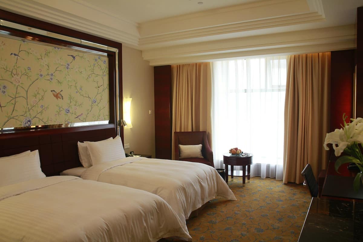 Ein Hotelzimmer mit zwei Betten, einem großen Blumenbild an der Wand, einem Schreibtisch mit Lampe und Blick ins Freie durch große Fenster.