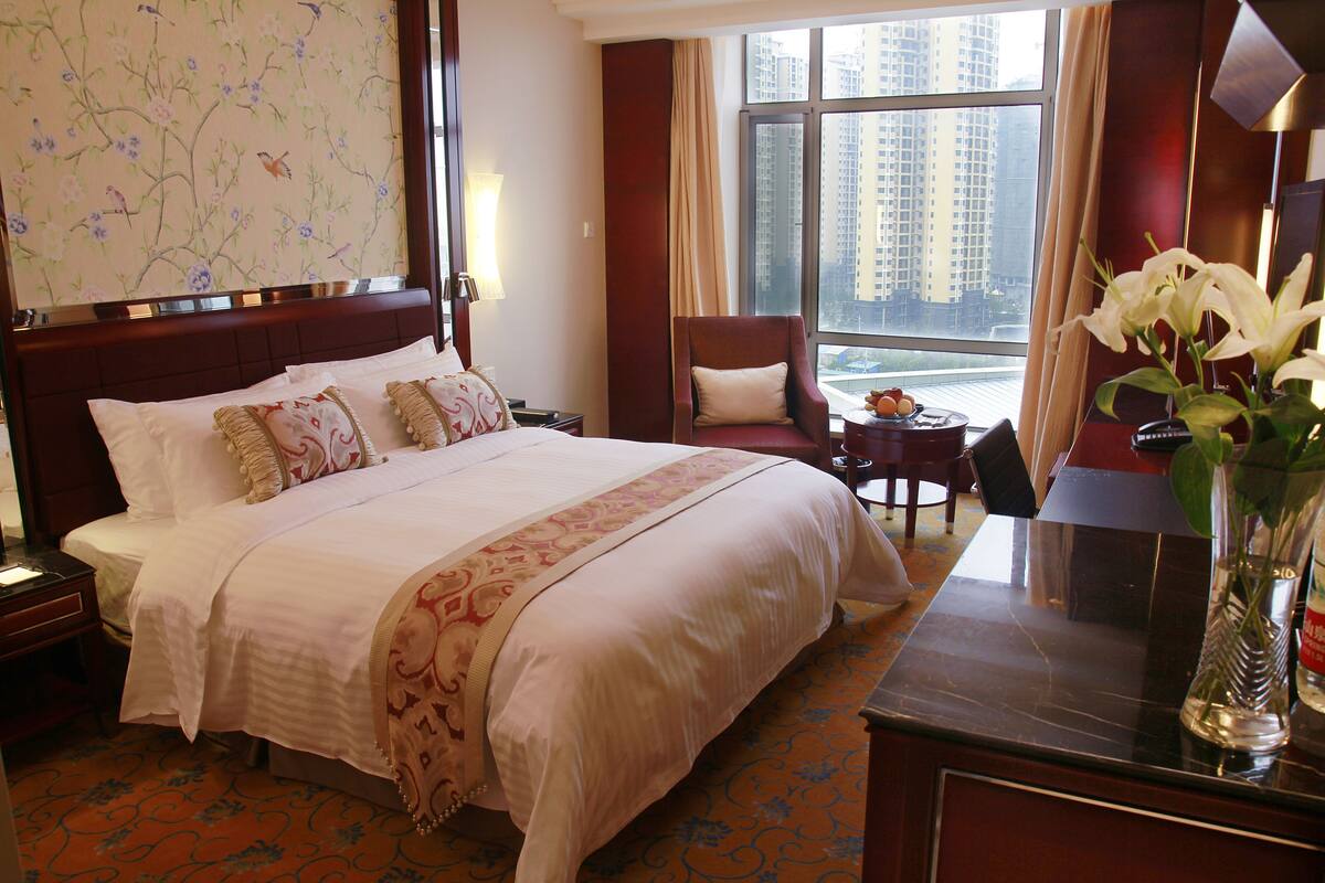 Ein Hotelzimmer mit einem großen Bett, einem Schreibtisch, einem Sessel, einer Vase mit Blumen und Blick auf hohe Gebäude durch das Fenster.