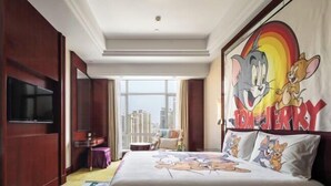 Habitación de hotel con una colcha grande de temática de dibujos animados, un televisor y vistas de la ciudad.