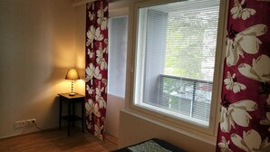 A room with a wooden floor, a small table with a lamp, a window with blinds, and a floral curtain.
