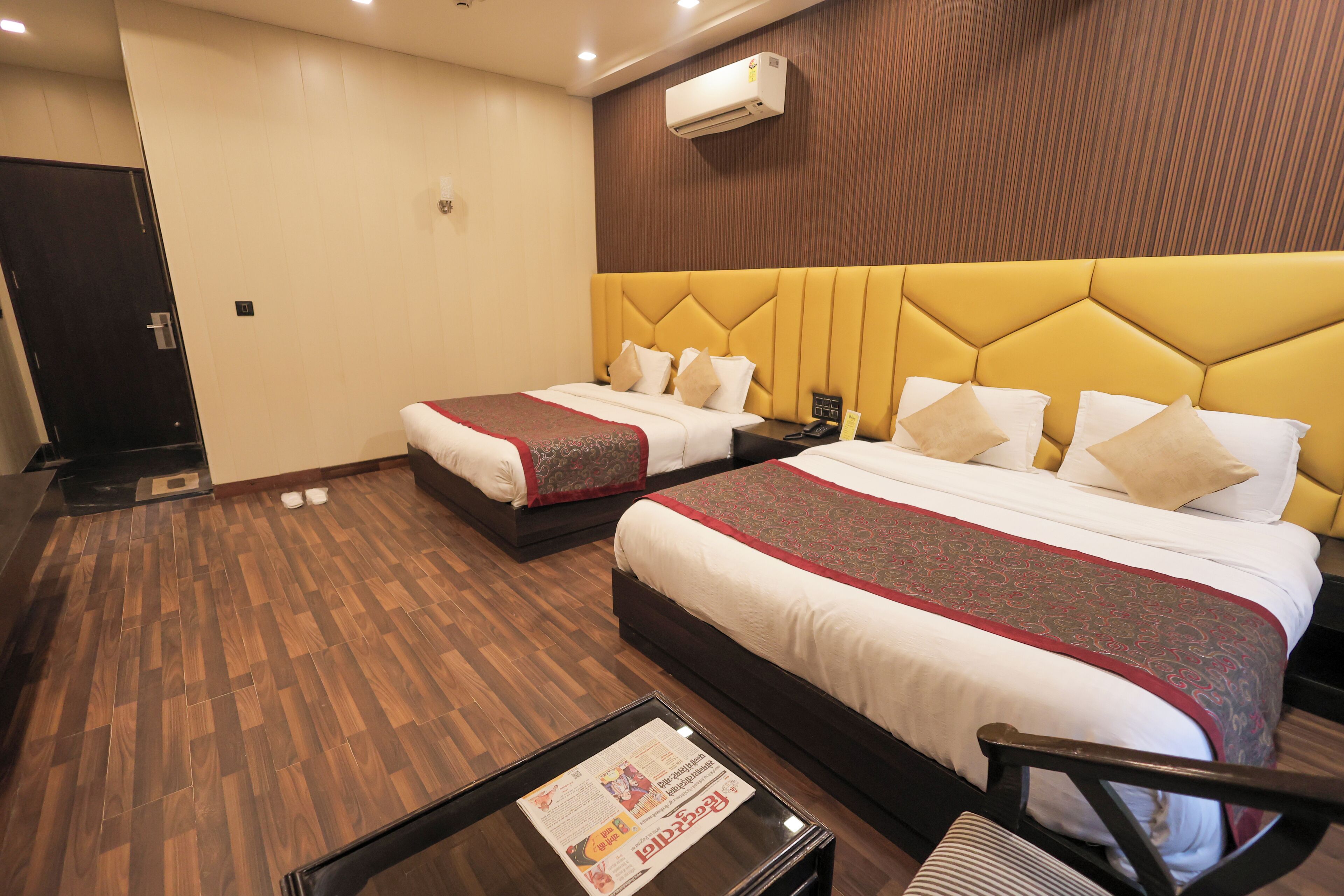 A hotel room with two beds, a wooden floor, a small table with a newspaper, and an air conditioning unit on the wall.