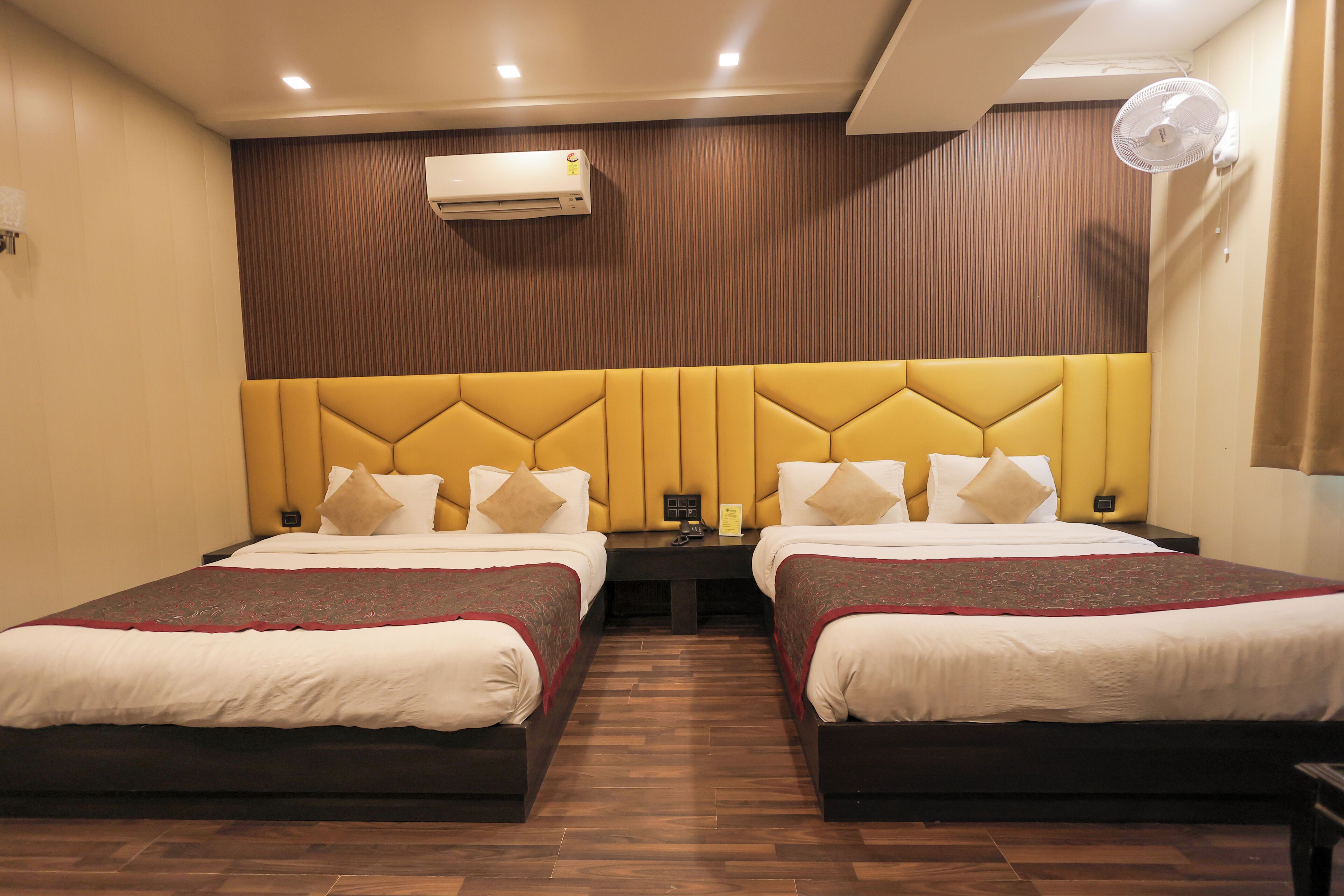 A hotel room with two beds, a wooden headboard, an air conditioning unit, a bedside table with a lamp, and a wall-mounted fan.
