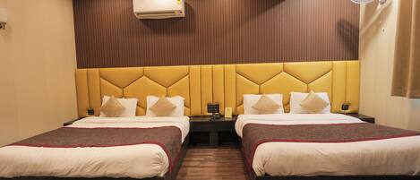 A hotel room with two beds, a wooden headboard, an air conditioning unit, a bedside table with a lamp, and a wall-mounted fan.