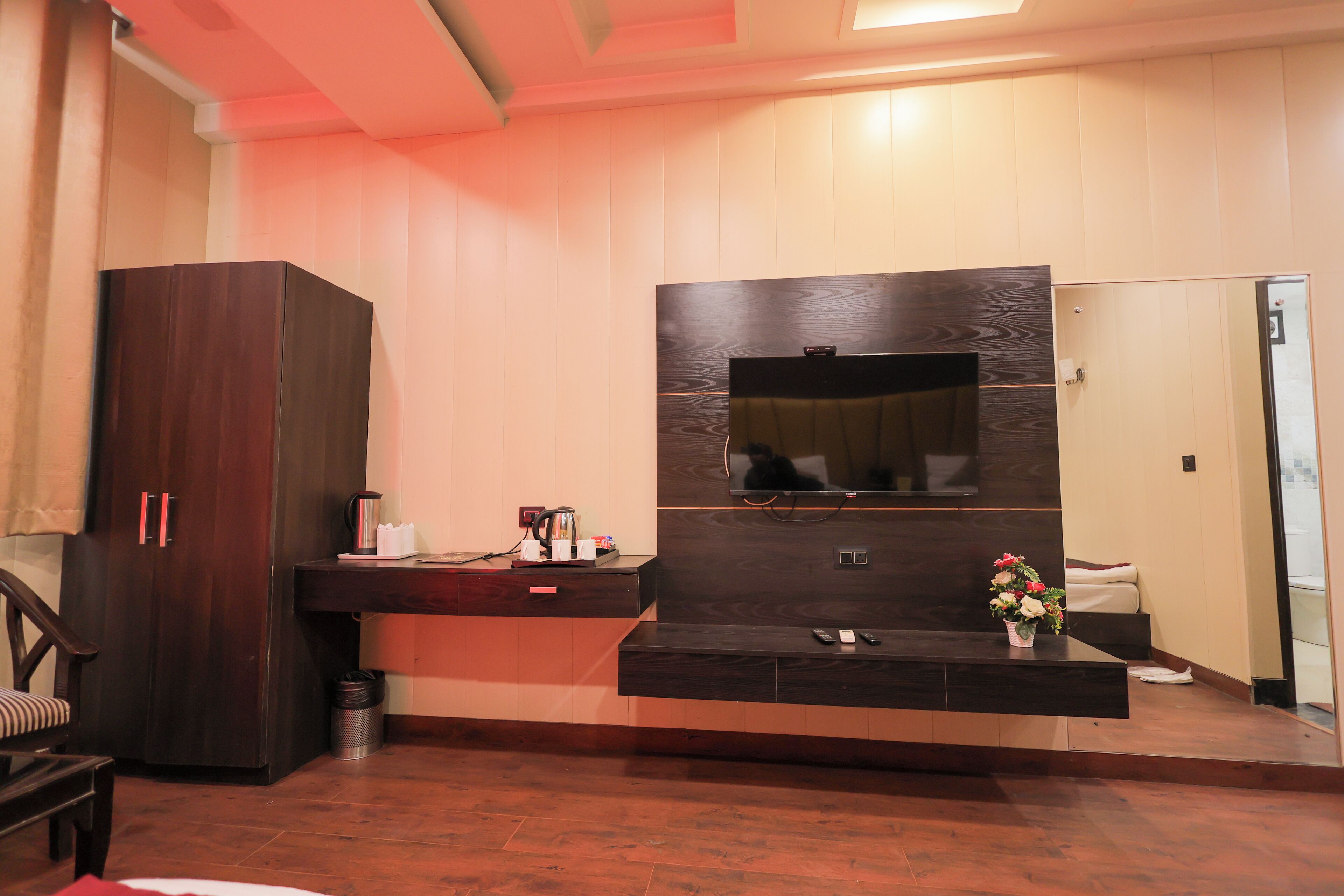 A modern room with a dark wood wardrobe, a flat-screen TV mounted on a wooden wall, a small desk with a coffee maker, and a flower vase.