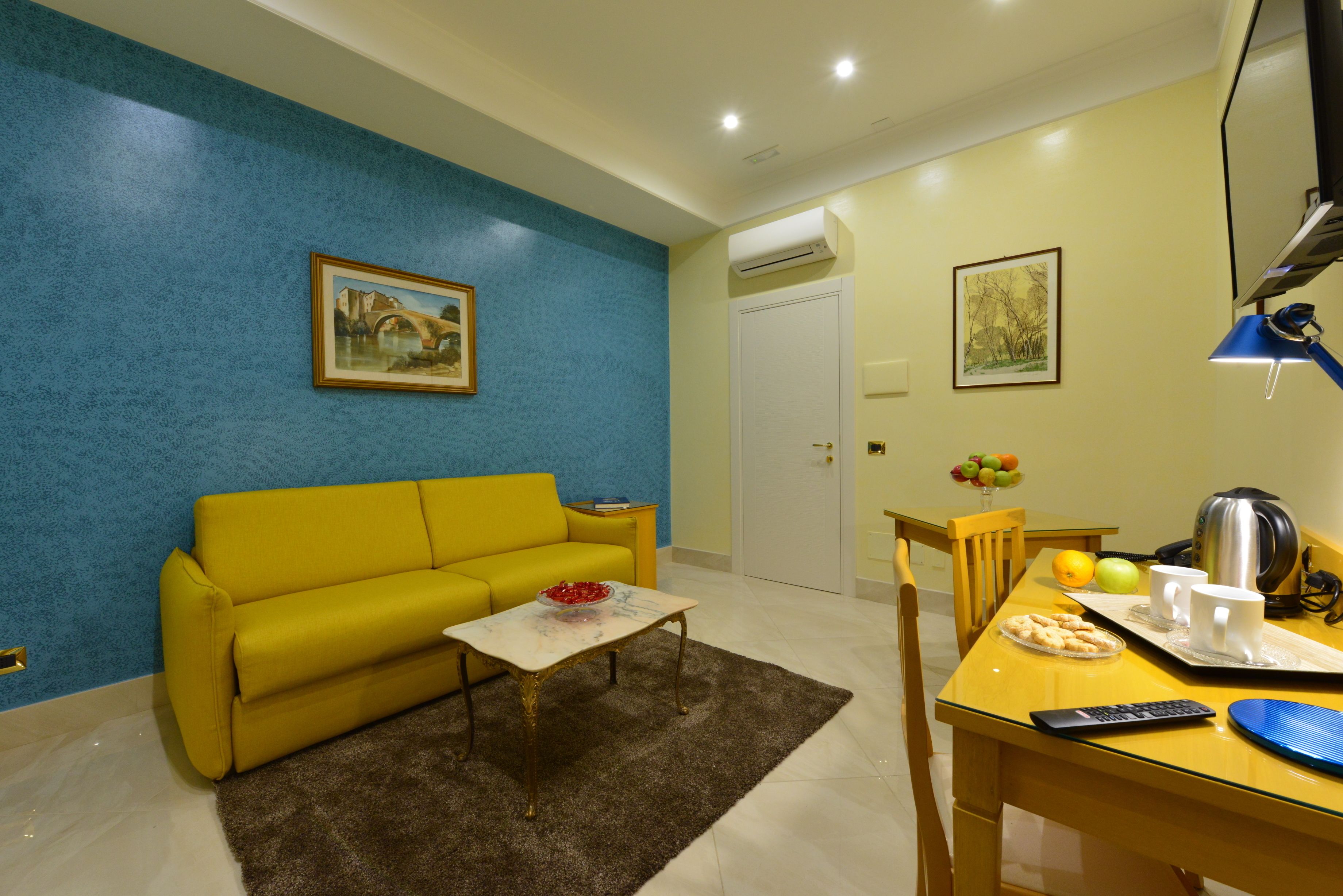 Un salón con un sofá amarillo, una mesa de centro, una mesa de comedor con sillas, un televisor y obras de arte en las paredes.