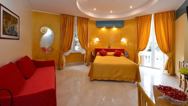 Una habitación de hotel con cama, sofá rojo, espejo y ventana con cortinas.